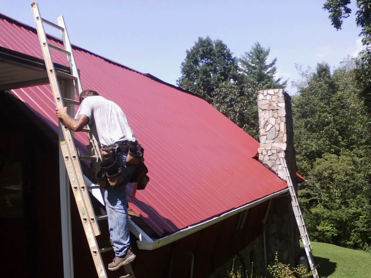 Roofer on a ladder beside a red metal roof and stone chimney — Soffit & Fascia Repair by Roofing Brothers in Bernville